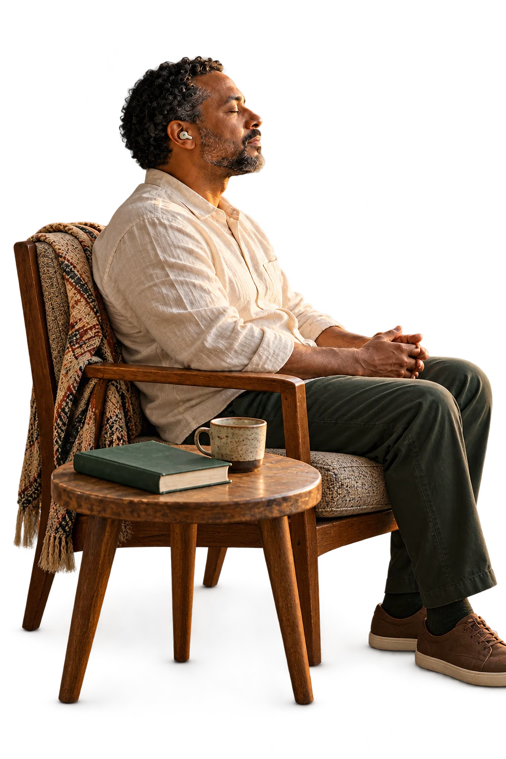 A man wearing an earbud sits peacefully in an armchair beside a table with a book and mug.
